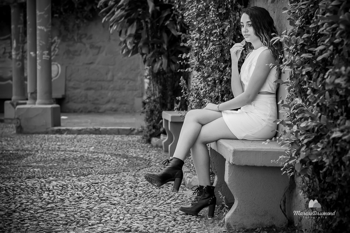 Fotografia preto e branco onde a modelo está sentada em um banco e segurando o brinco da sua orelha direita e com a mão esquerda no joelho com as pernas cruzadas olhando para frente em uma praça