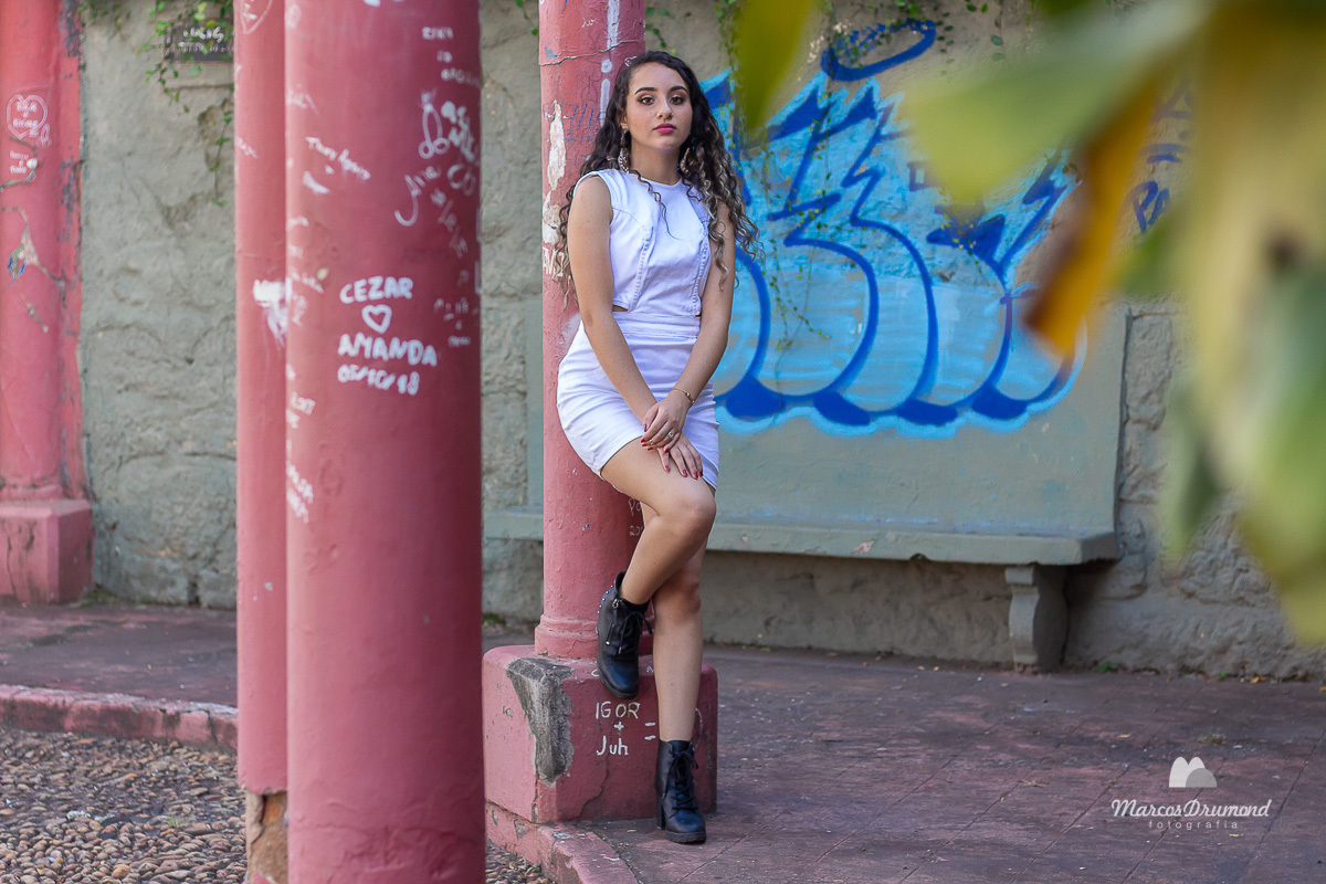 Fotografia de uma modelo de corpo inteiro, onde ela está usando um vestido na cor branco e uma bota na cor preta, a modelo está colocando as mãos no joelho direito e olhando para frente 