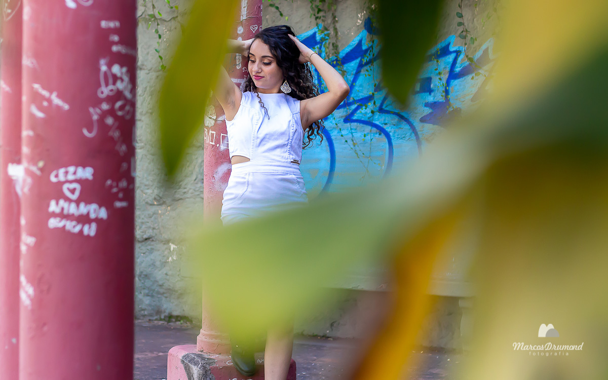 Fotografia da modelo Karoline feita em Itabira/MG onde a mesma está usando um vestido jeans na cor branco e uma bota na cor preta, cabelos longo e cacheados e brincos longos, a modelo está segurando o cabelo na hora do ensaio fotográfico