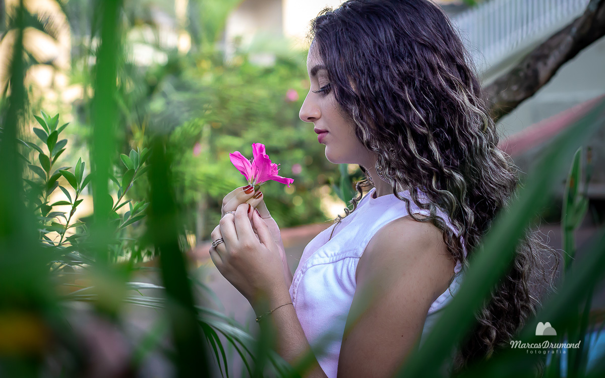 Ensaio fetográfico realizado na cidade de Itabira/MG onde a modelo está usando  uma vestido jeans na cor branco e segurando uma flor e olhando para ela entre as folhas de uma vegetação 