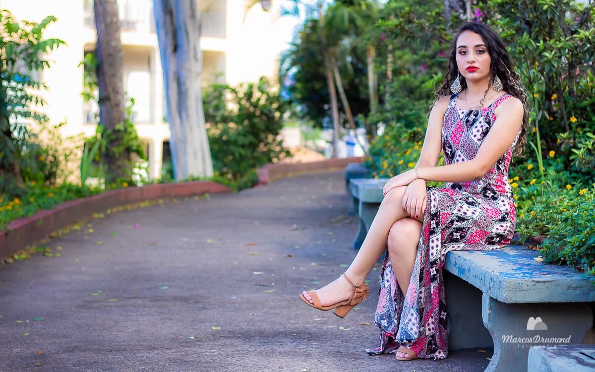 Registro fotográfico feito de um ensaio fotográfico feminino na cidade de Itabira/MG, onde a modelo está sentada em m banco na praça e usando um vestido florido longo com um par de sandália com as mãos no joelho, batom vermelho olhando para câmera
