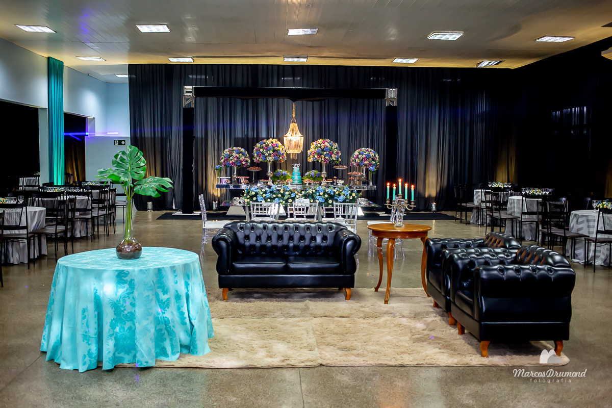 Fotografia colorida da decoração da festa da debutante Natielle Cristina realizado na cidade de Itabira/MG onde há um mesa com um pano na cor azul e flores na cor verde sobre a mesma, sofás na cor preto, mesa de madeira, e a mesa de bolo no funco decorada