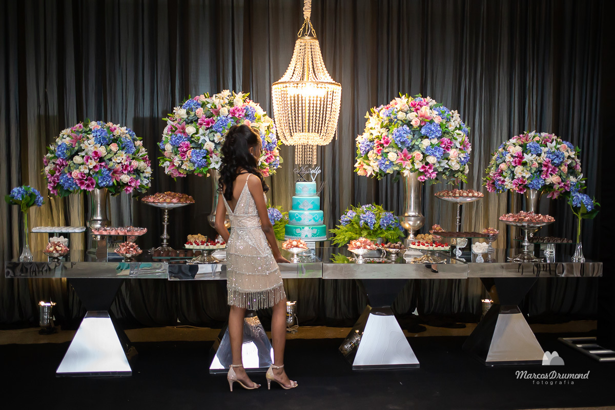 Fotografia colorida de aniversário de 15 anos de Natielle Cristina na cIdade de Itabira/MG, onde a debutante está de costas olhando para mesa com as mãos sobre a mesma, ela está com um vestido na altura do joelho e sandália cabelos pretos