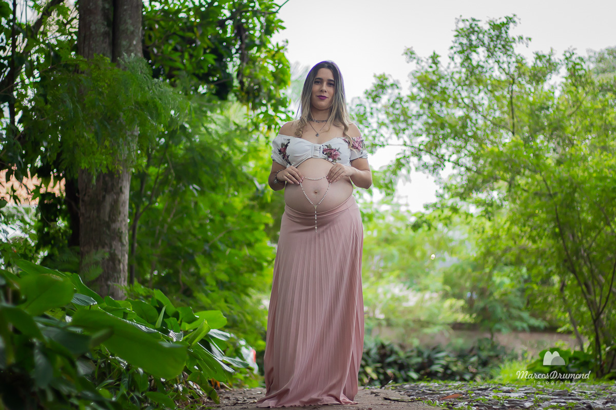 Ensaio gestante realizado na cidade de Itabira/MG, onde a gestante está com uma saia longa na cor clara, e com um terço fazendo um coração na barriga, andando de frente para a câmera e usando um top na cor branco, na região há muita vegetação verde