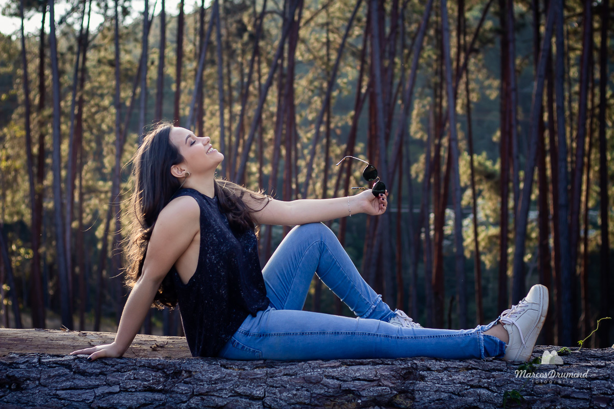 Fotografia colorida de uma de debutante sentada em uma árvore usando uma calça jeans e blisa sem mangas com a mão no joelho olhando para cima e com os óculos na mão com os cabelos pretos e longos pousando para o ensaio fotográfico em comemoração de