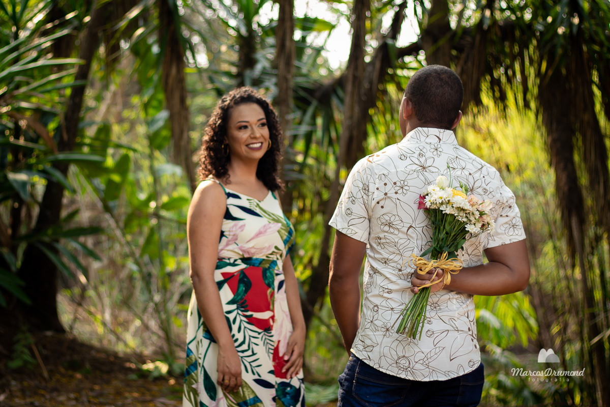 Fotografia de pré casamento onde a noiva está vestida com um vestido florido olhando para o noivo e ele de frente para ela segurando um buquê de rosa, ele está vestido do uma camisa social clara