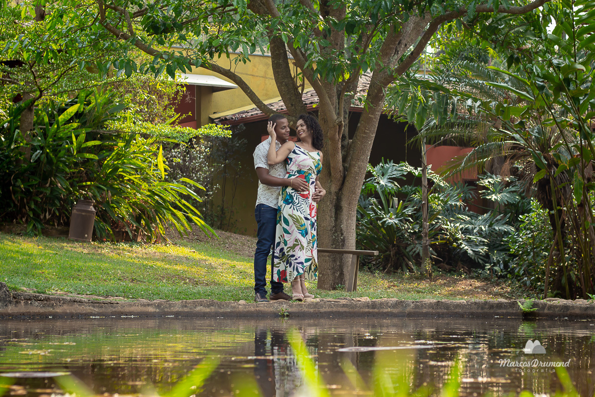 Fotografia de casal de corpo inteiro onde o casal está de frente para um lago e ela está de costa para ele com a mão direita no rosto dele e ele com as mãos na cintura dela e ambos sorrindo na foto do sítio que fizeram as fotos de pré casamento em Itabira