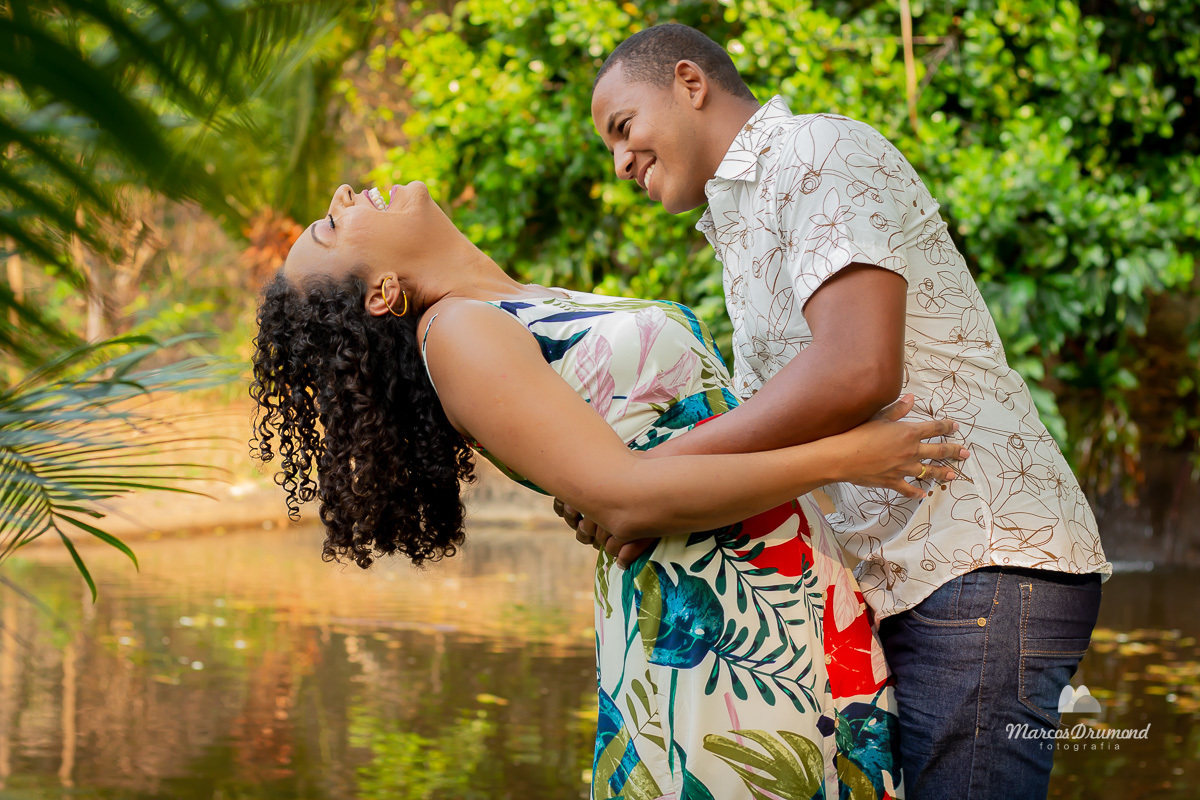 Fotografia colorida de um casal em pré casamento onde o casal está de frente para uma lagoa e ele está segurando ela onde ela está inclinado para trás onde ambos estão sorrindo para as fotos de pre wedding no sítio que escolheram para fotografar