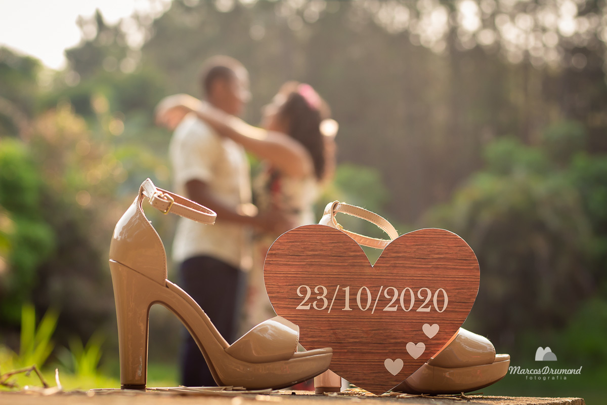 Foto de um casal em pre casamento com um coração com a data do casamento deles, onde este coração está encostado na sandália e o casal está desfocado no fundo da foto de frente um para o outro e uma paisagem linda no fundo com um sol colorido e raios 