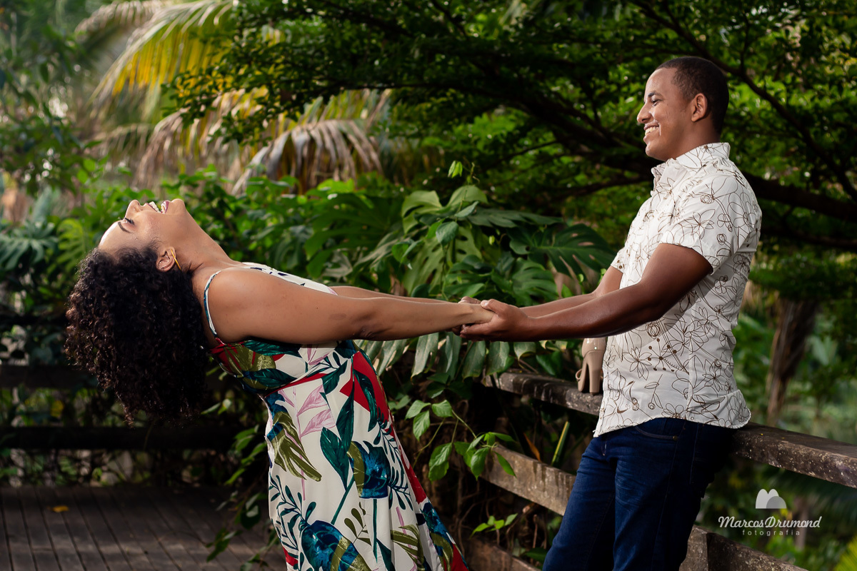 Fotografia de pré casamento onde os noivos estão de frente um para o outro o noivo está escorado em uma cerca segurando as mãos da noiva e ela está com o corpo inclinado para trás sorrindo para ser fotografada, o noivo também está sorrindo e há uma linda 