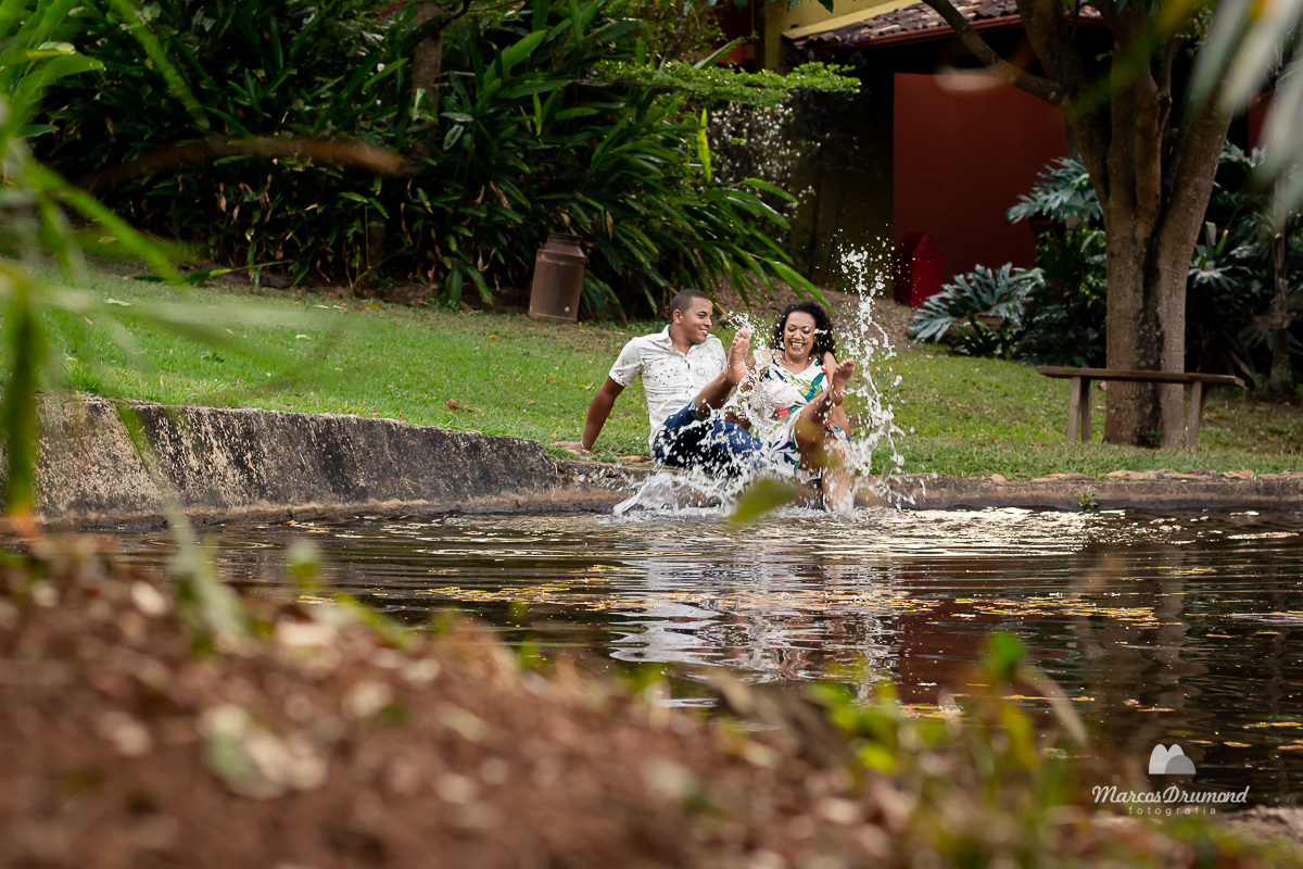 Fotografia colorida de pre casamento onde o casal está sentado na beira de uma lagoa brincando de jogar água com o pés e com as mãos para trás, eles estão sorrindo e se divertindo para as fotos