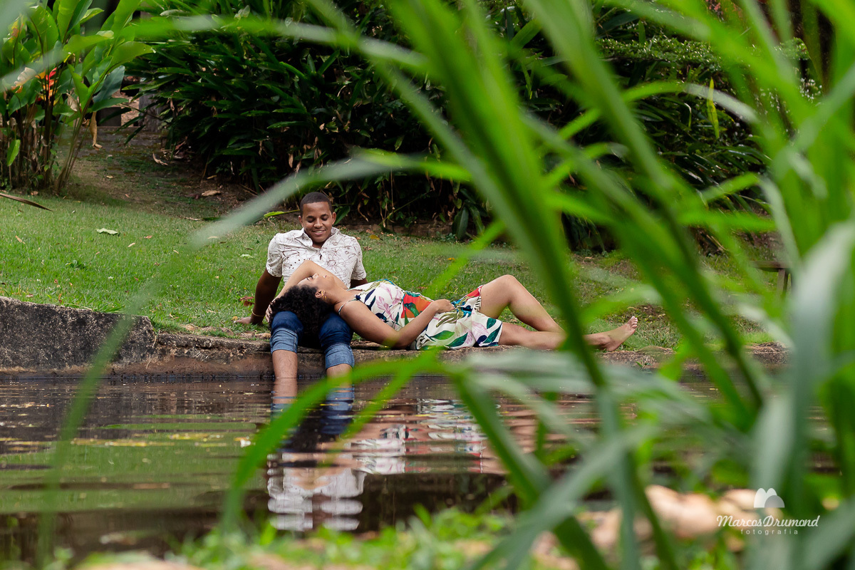 Fotografia de pré casamento onde a noiva está sentada na beira de uma lagoa e a noiva está deitada no colo do noivo, a foto foi feita atrás de uma moita de ramos verdes super bacana