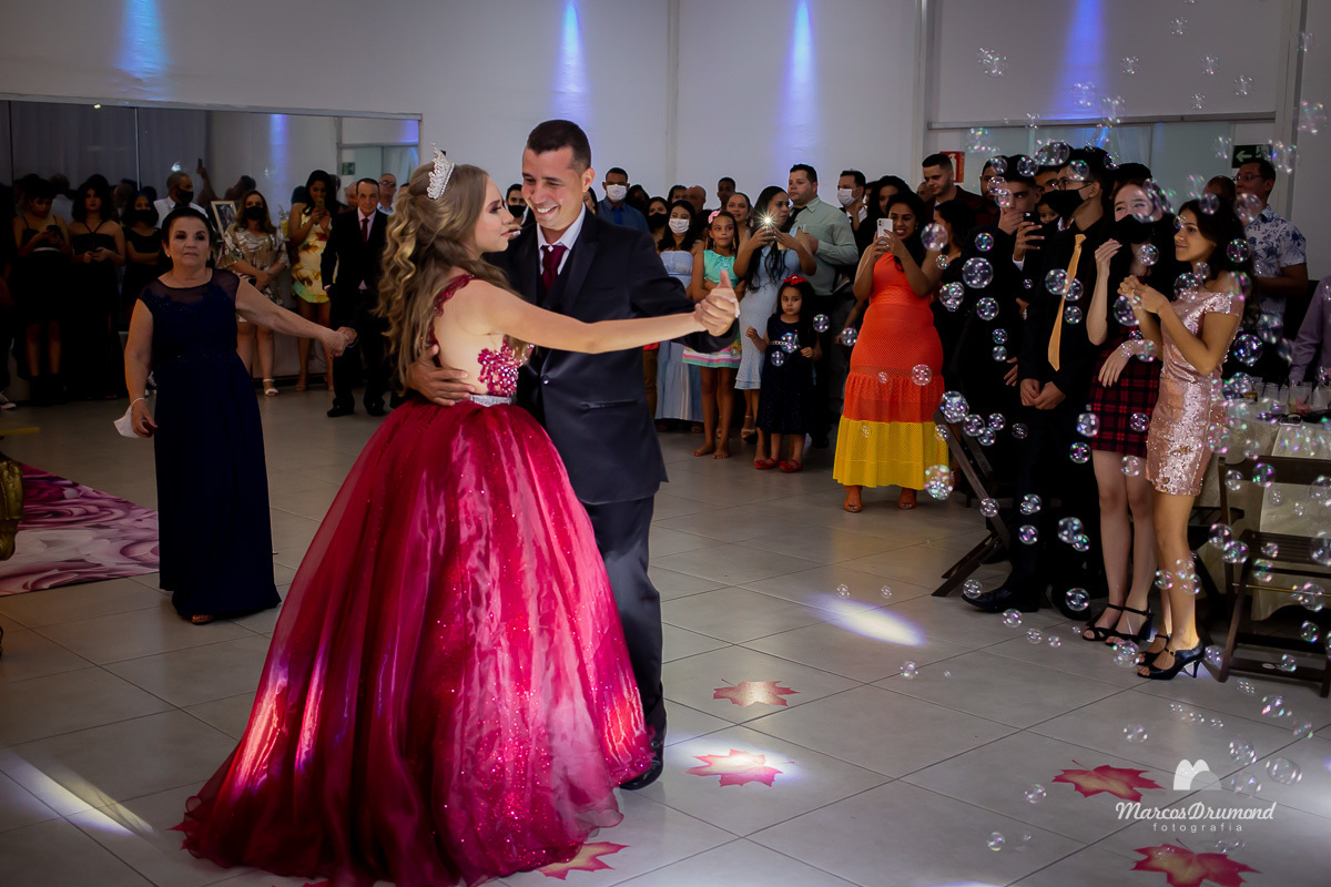 Debutante loira, cabelos longo com arco branco na cabeça usando vestido vermelho, dançando com o pai que está d eterno preto, camisa branca e gravata vermelha, com os convidados olhando e fotografando com o celular em sua festa de 15 anos