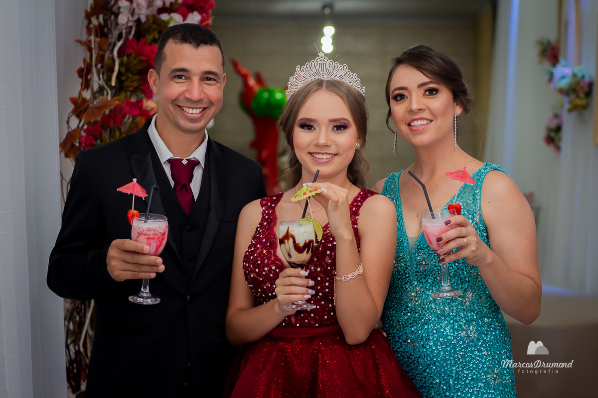 Fotografia de família no aniversário de 15 anos de Julia Mascarenhas, onde a mesma está usando um vestido vermelho tomando um brink com a mãe que está usando vestido verde e segurando um copo com bebida ao lado do pai que também está bebendo e sorrindo