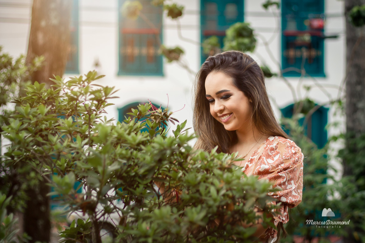 Fotografia de quinze (15) anos onde a debutante está próximo de umas flores sorrindo e olhando para as flores, o local é urbano e atrás dela tem um casarão antigo na cidade de Itabira/MG. Ela está usando blusa estampada, bermuda branca e rasteirinha.