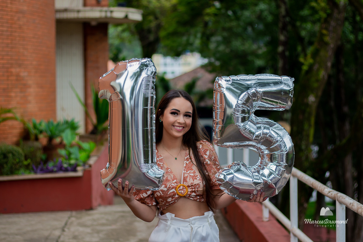 Fotografia de ensaio de debutante segurando o número quinze (15) urbano onde ela está com uma short branco e blusa estampada aberta do peito, usando um cordão, olhando para frente e sorrindo, ela tem cabelos longos e pretos, está em área urbana no jardim.