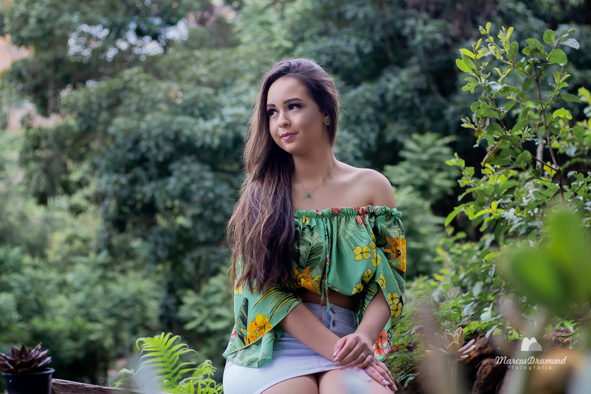 Fotografia de 15 anos onde a modelo, debutante está usando uma blusa verde tomara que caia florida e saia branca sentada em um banco com as pernas cruzadas e as mãos uma sobre a outra olhando para o lado, com o cabelos longos próximo de uma vegetação flor