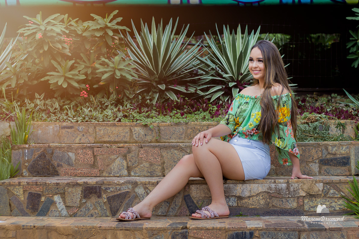 Ensaio de quinze anos onde a debutante está com uma blusa tomara que caia verde florida e saia branca de rasteirinha e cabelos longos sentada em uma escada com uma perna dobrada, cabelos longos, olhando e sorrindo, uma mão está apoiando no chão e a outra 