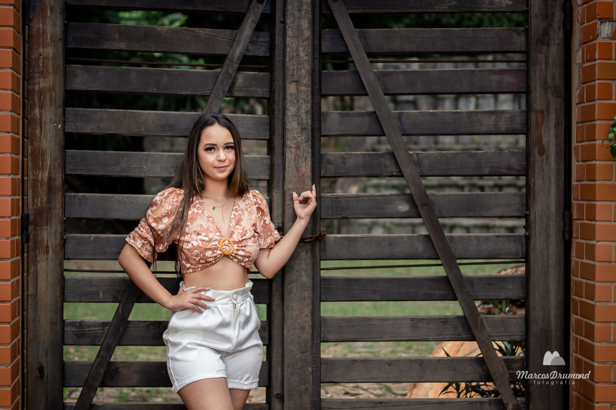 Fotografia de debutante na cidade de Itabira/MG no parque da Água Santa, onde a debutante está próximo ao portão de acesso ao parque e usando uma blusa florida marrom claro e uma bermuda short branco com uma rasteirinha e um cordão, com os cabelos longos 
