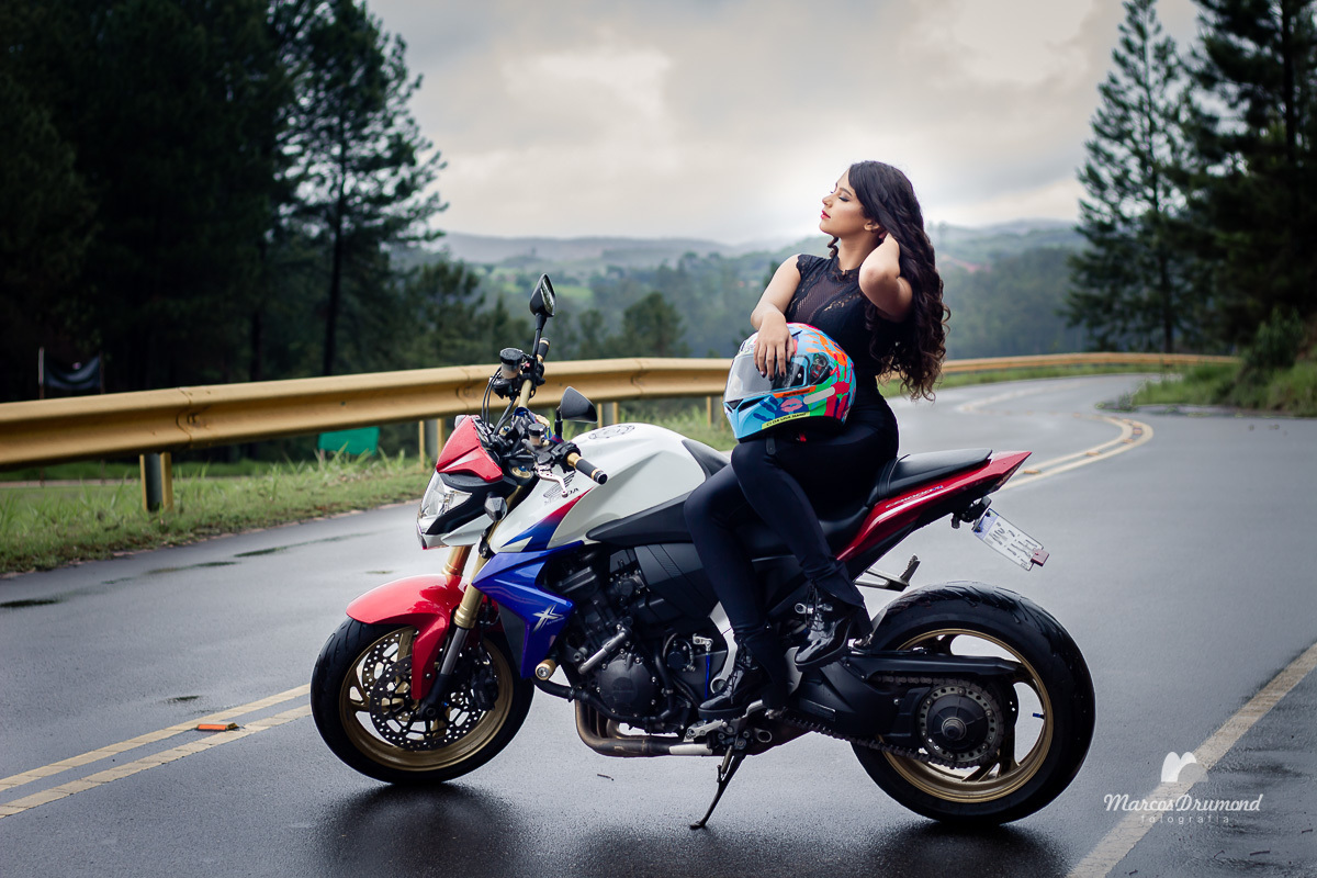 Ensaio modelo 15 anos com moto no asfalto, onde a debutante está com as mãos no cabelo, sentada na moto com as mãos no capacete e o dia chuvoso, mas a debutante está fotografando mesmo assim em uma moto branca, foto linda de debutante