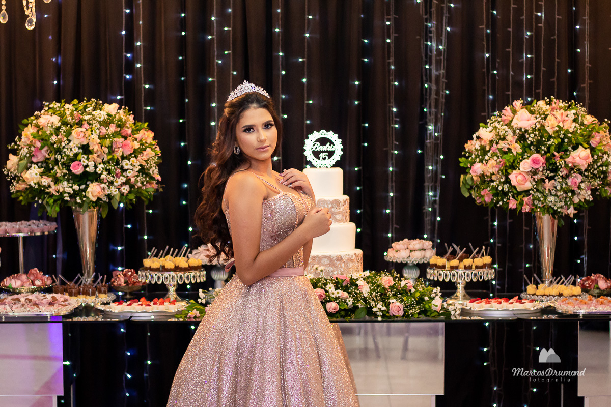 Fotografia de 15 anos da debutante Beatriz em Itabira MG, onde a mesma está cum as mãos próximo ao  peito, olhando para o lado e usando brincos e coroa no cabelo próximo a mesa de bolo em sua festa, assim a mesma está comemorando muito feliz
