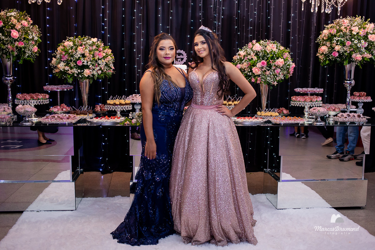 Fotografia de uma debutante em sua festa de 15 anos com sua mãe, onde a mesma está com um vestido longo na cor bege próximo a mesa de bolo, com os cabelos longos e pretos, usando brincos brancos e coroa no cabelo, com uma das mãos na cintura junto a mãe