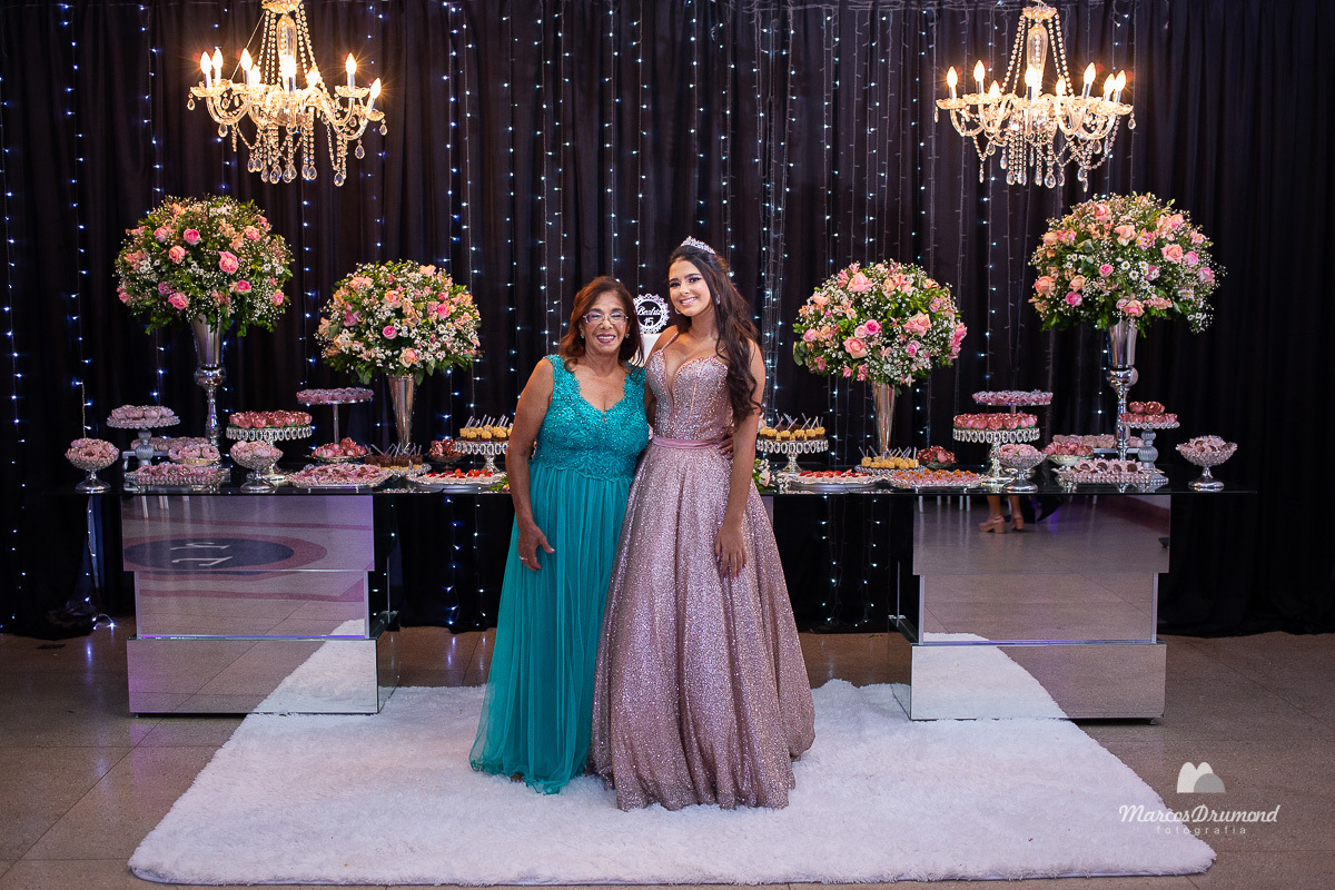 Foto de uma menina comemorando os seus 15 anos em seu aniversário, onde ela está com um vestido longo cor creme e cabelos pretos e longo, junto a sua avó que está usando um vestido na cor azul e ambas estão com as mães na cintura uma da outra olhando para