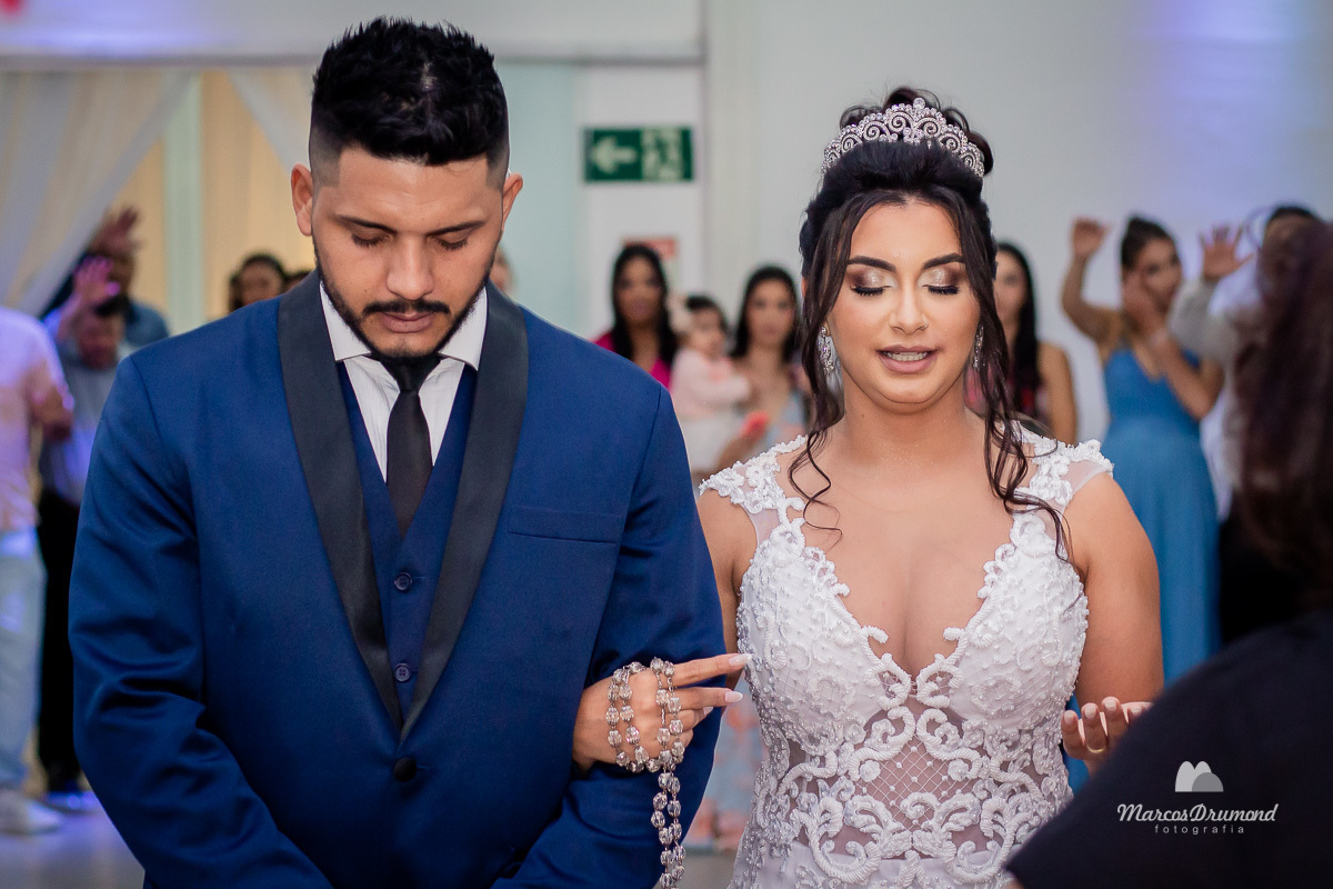 Casamento de Carla e Luan, onde a noiva está com o braço no noivo e ela está usando uma vestido branco com um terço na mão e o noivo está usando um blazer na cor azul, ambos os noivos estão olhando para baixo com os olhos fechados