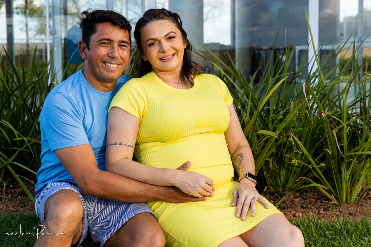 homem sentado abraçando esposa grávida em ensaio no Ybi condomínio em natal rn na primeira gravidez em ensaio externo no campo de fotografia de grávida
