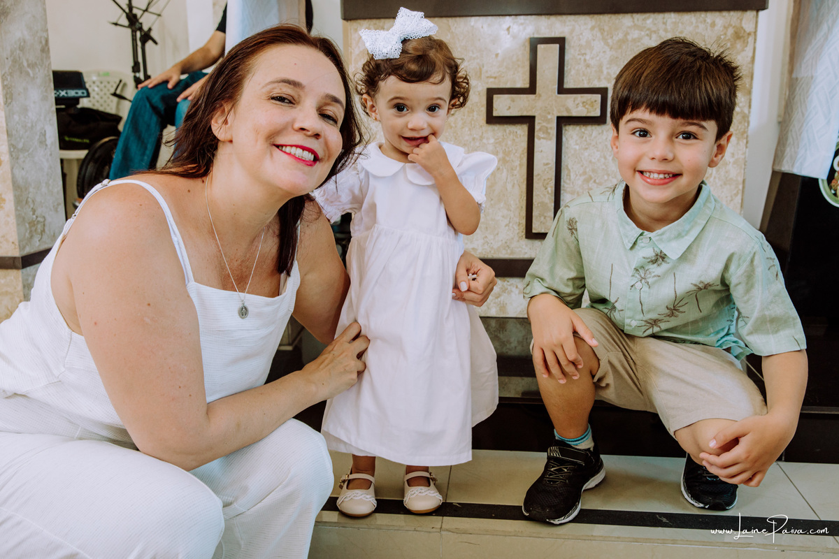 Criança em pé e sorrindo posando para foto ao lado de primo e mãe em seu batizado