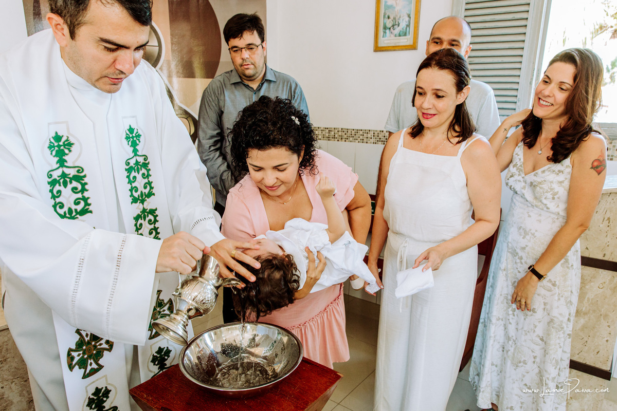 criança no colo da madrinha ao lado de pais e padrinhos enquanto padre a batiza com agua benta no seu batizado