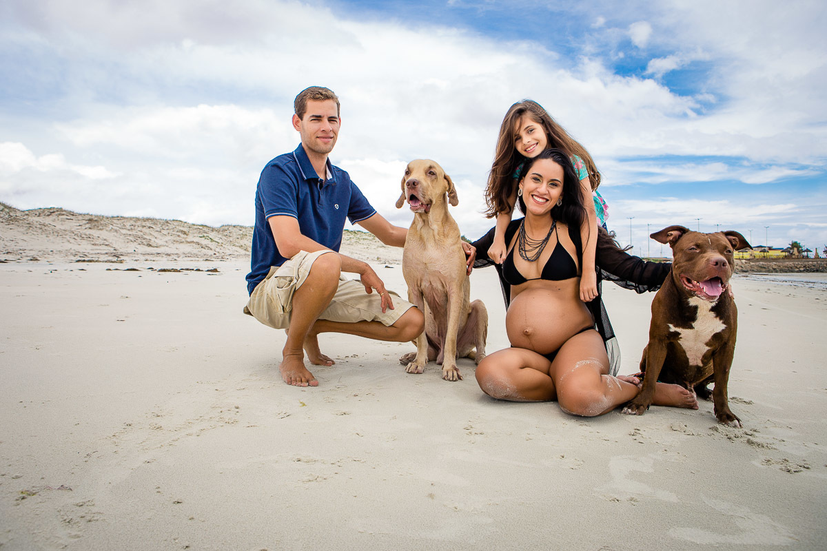gestante com marido, enteada e cachorros sentados no chão da praia