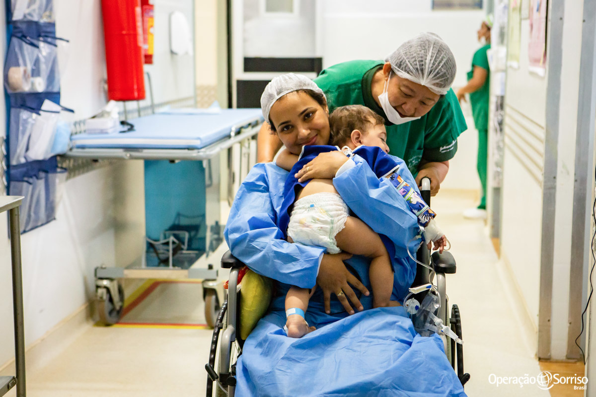 mãe sendo carreganda em cadeira de rodas do centro cirúrgico com filho após a cirurgia