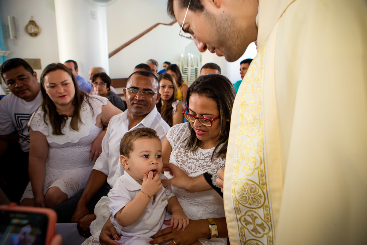 criança olhando para a o padre enquanto ele faz sinal da cruz em sua testa e pais e padrinhos o olhando