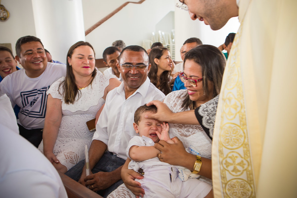 criança se esquivando do padre quando ele faz sinal da cruz na sua testa e padrinho e pais sorrindo