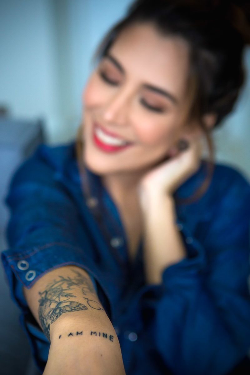 mulher sentada ao chão encostada no sofá com mão no rosto e mostrando tatuagem I am Mine e usando camisa de botão jeans