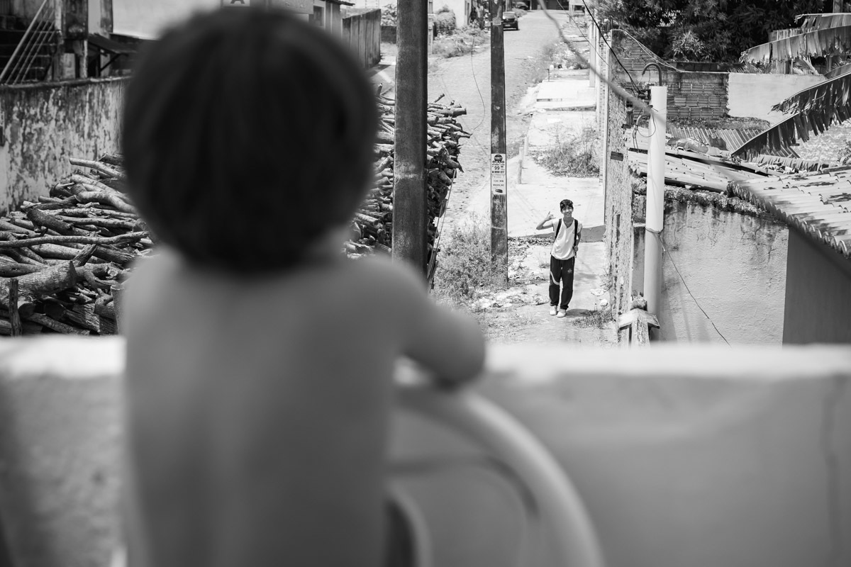 criança em cima da cadeira e encostada no muro olhando para o irmão que vem da escola