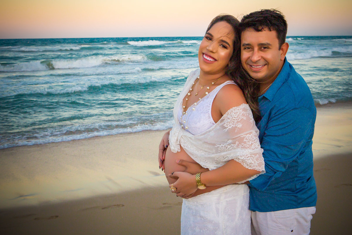 Gestante e marido abraçados na praia com mão na barriga