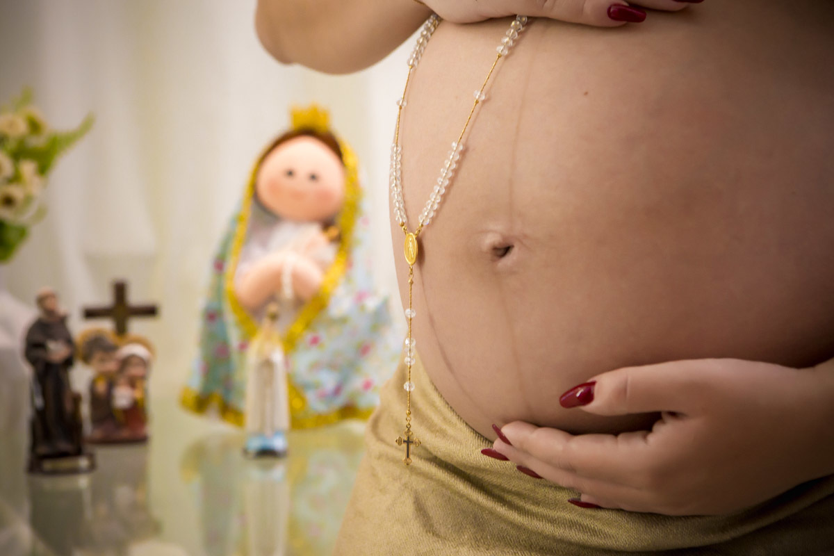 Gestante segurando terço na barriga com santos ao fundo