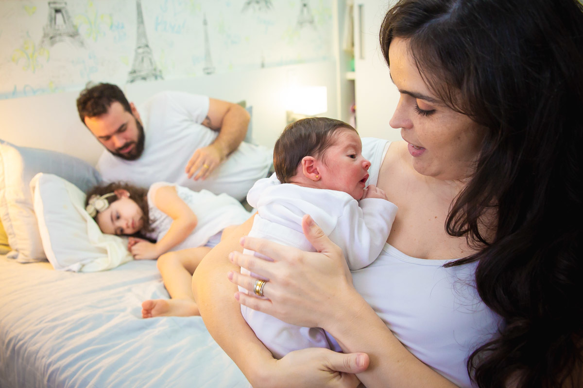 mãe segunda bebe no colo e irmã mais velha deitada na cama olhando pra baixo chateada, com ciúmes e pai conversando com ela.