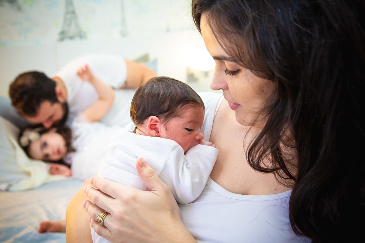 mãe segunda bebe no colo e irmã mais velha deitada na cama olhando pra elas com ciúmes e pai conversando com ela.