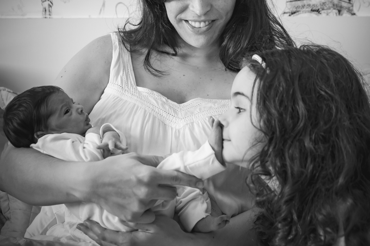 mãe segunda bebe no colo e irmã mais velha cheirando seu pezinho.