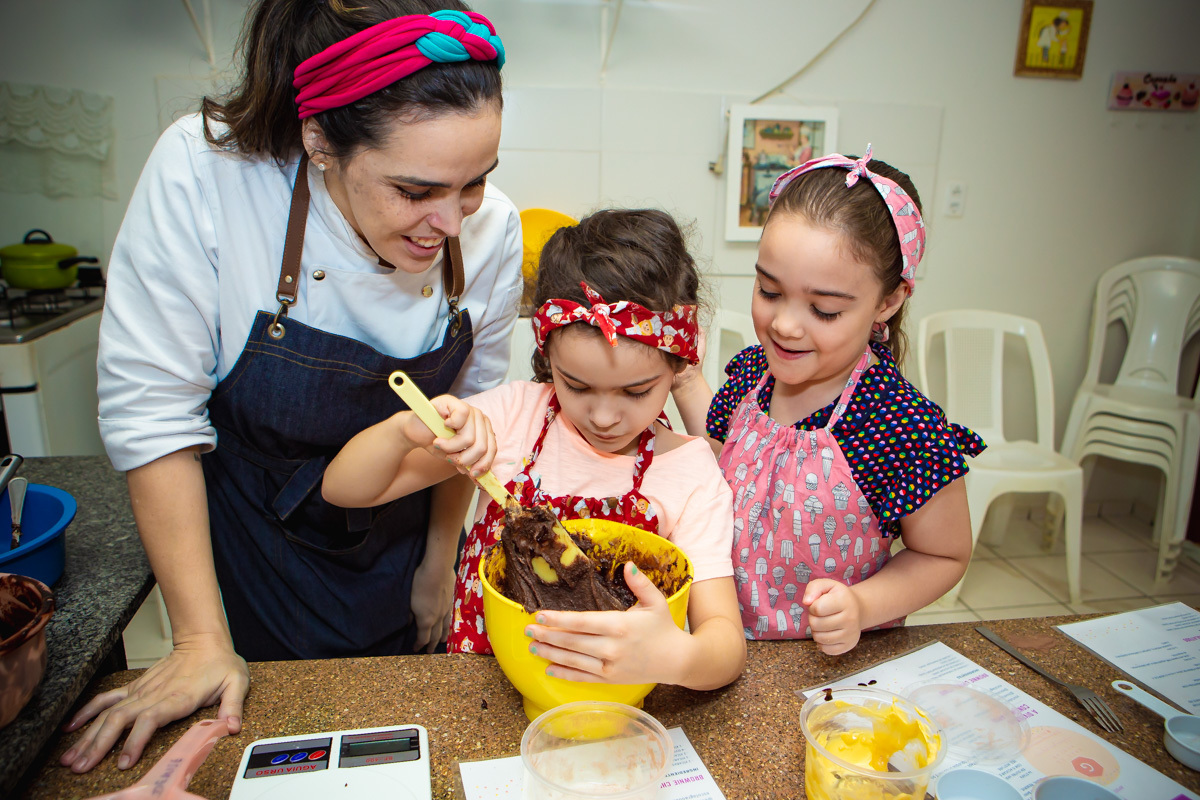 Menina batendo massa de brownie e chef olhando junto com outra criança 