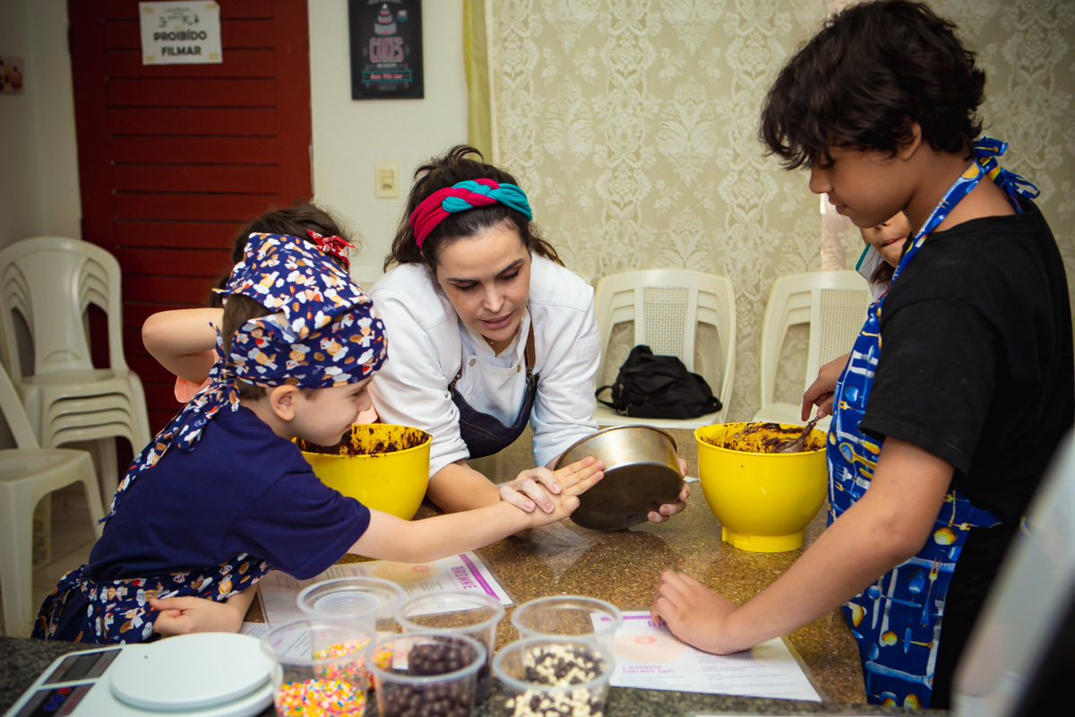 Chef mostrando pra crianças forma de bolo e quantidade que deve por de massa medindo com o dedo de aluno.