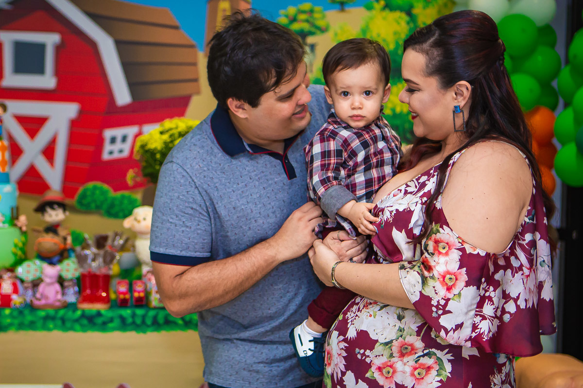 Pais olhando para aniversariante no colo em frente a mesa de bolo e doces do tema a fazendinha