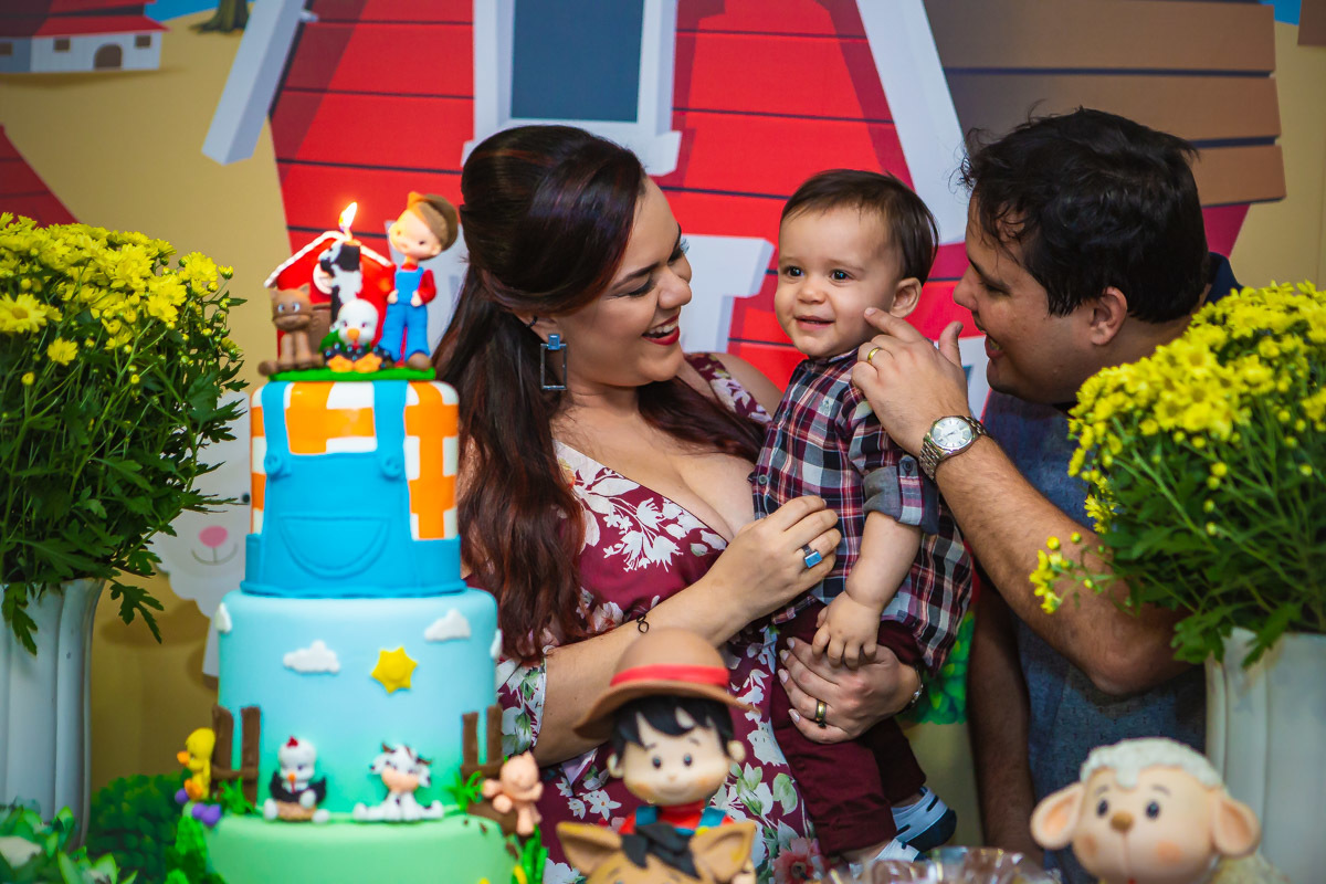 Pais olhando para aniversariante no colo e rindo em na hora do parabéns da festa com tema a fazendinha
