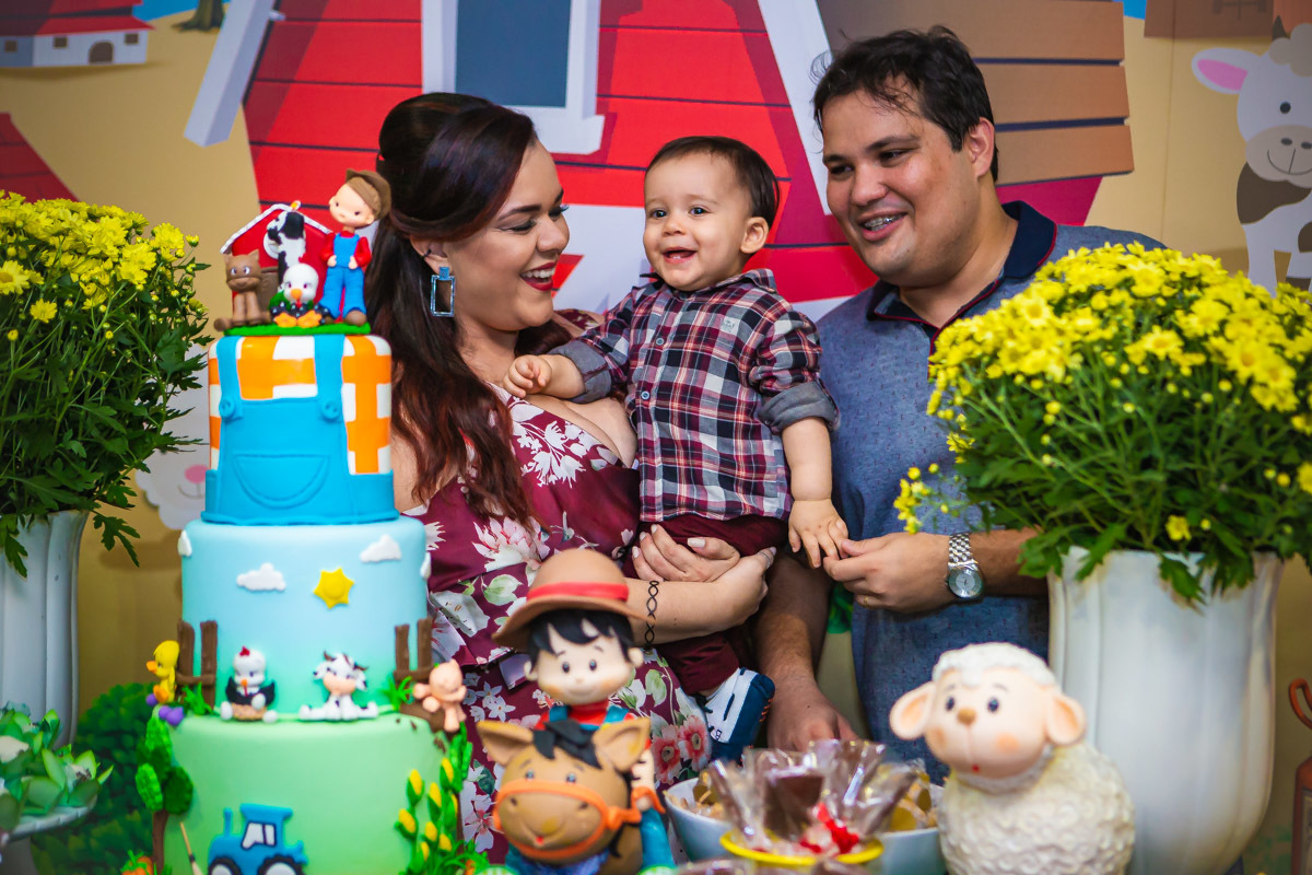 Pais olhando para aniversariante no colo e rindo em na hora do parabéns da festa com tema a fazendinha