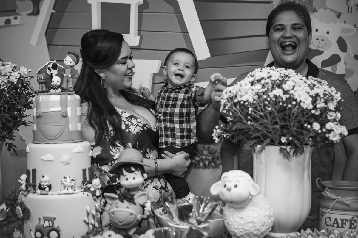 Pais olhando para aniversariante no colo e rindo em na hora do parabéns da festa com tema a fazendinha