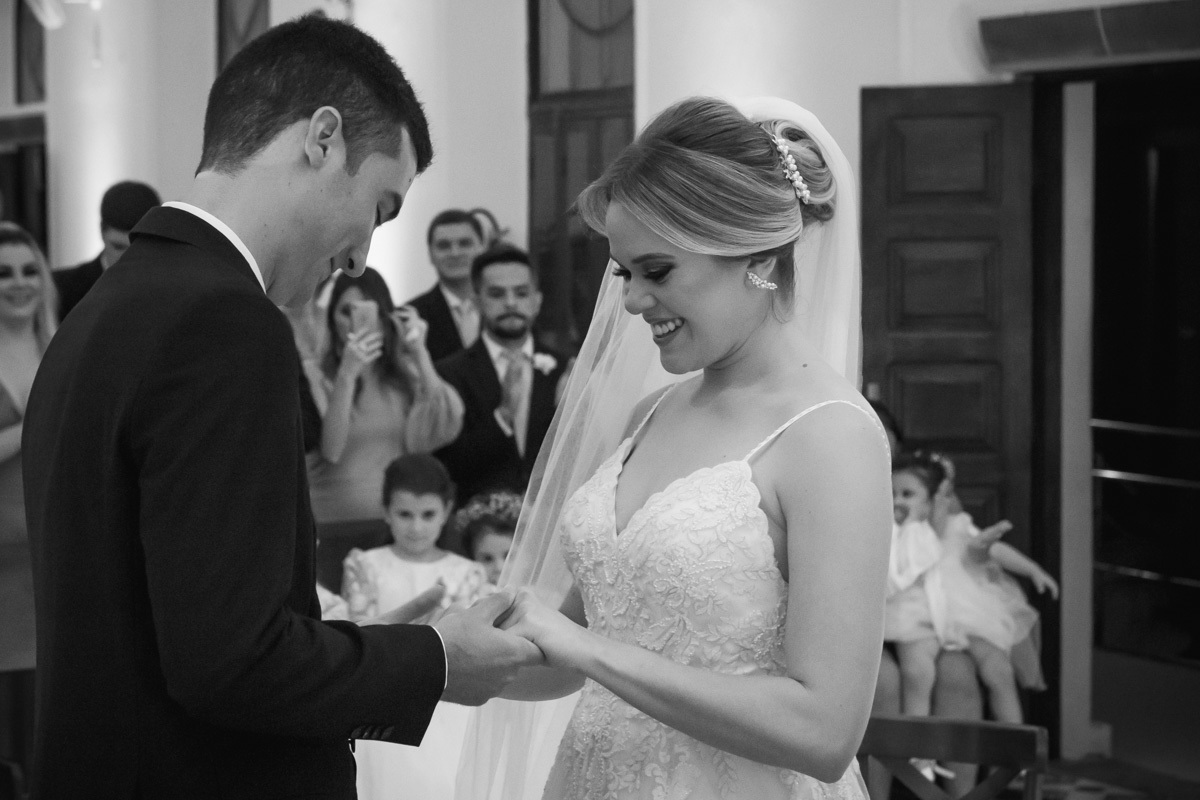 Celebração do casamento na Igreja Santa Terezinha, noivos olhando para as mãos e sorrindo enquanto fazem os rituais do casamento.