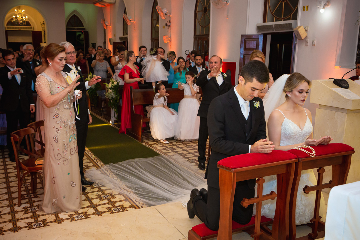 Celebração do casamento na Igreja Santa Terezinha, noivos ajoelhados recebendo benção de todos.
